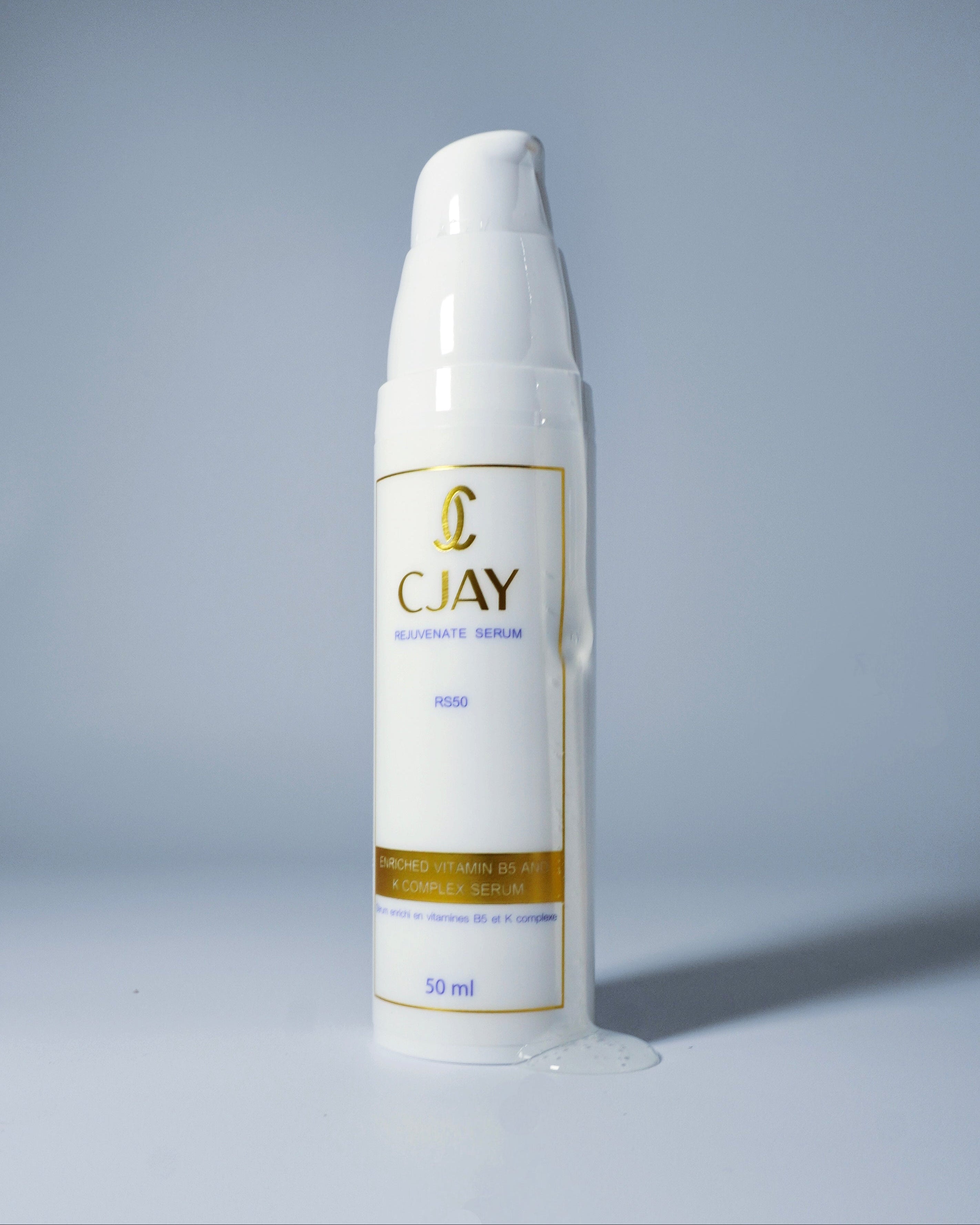 A white pump bottle of CJAY Rejuvenate Serum with gold lettering and borders, standing on a light gray surface against a matching gray background with a small puddle of serum at its base.