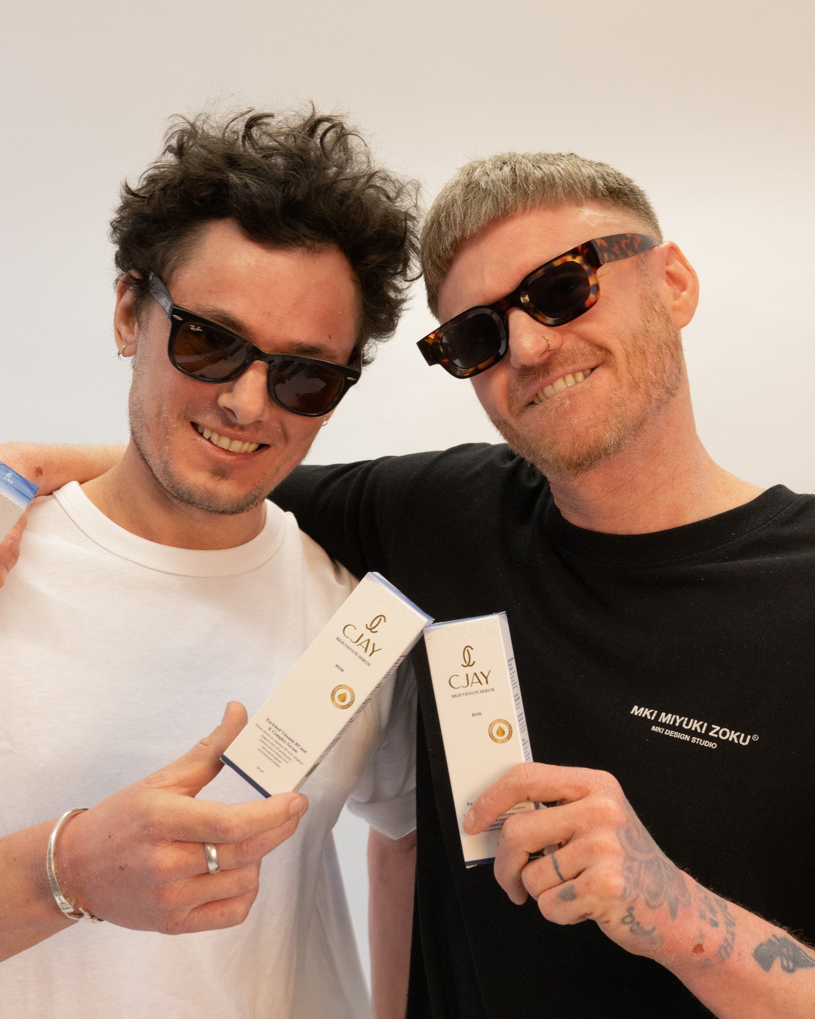 Two men wearing sunglasses and holding CJAY skincare products against a plain background